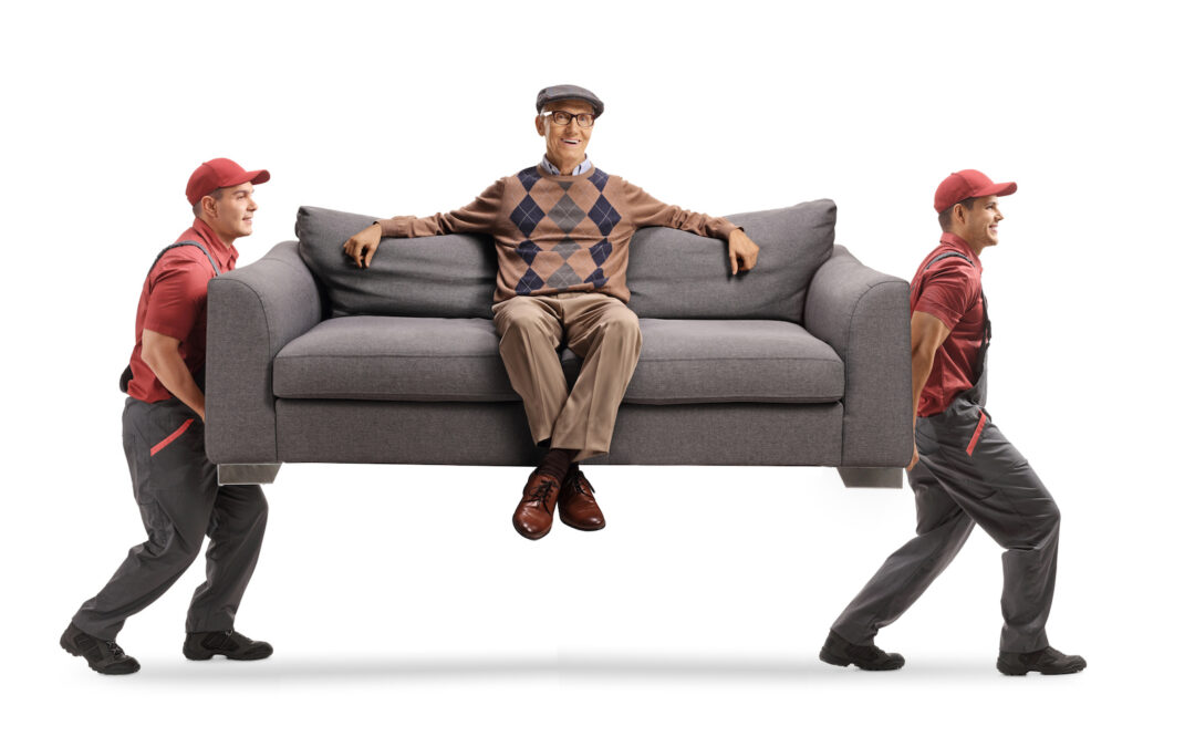 Movers Carrying A Sofa With An Elderly Man Resting Seated On The Sofa